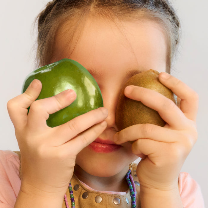 Young child holding a green apple and brown kiwi fruit over their eyes, wearing colorful beaded necklaces