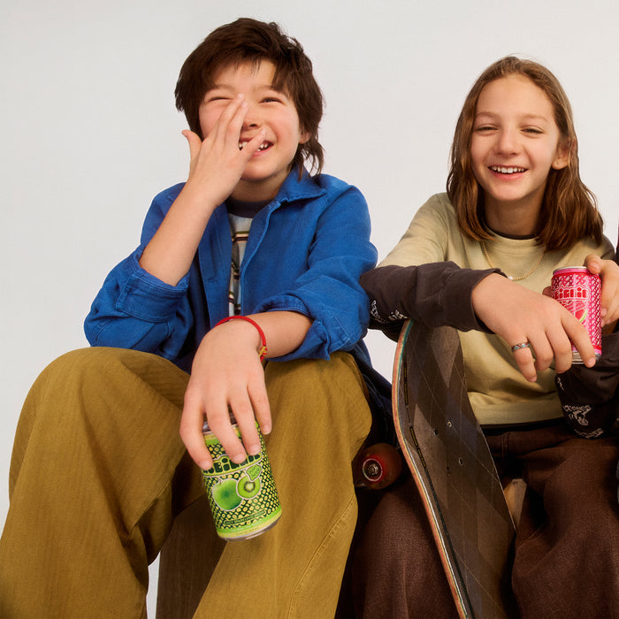 Two laughing teens sit together - one in blue jacket holding green can covering face, other in beige top holding red can with skateboard nearby