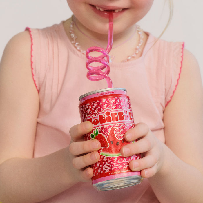 Child in pink dress drinking from red "Strawberry Watermelon" sparkling drink can through pink curly straw