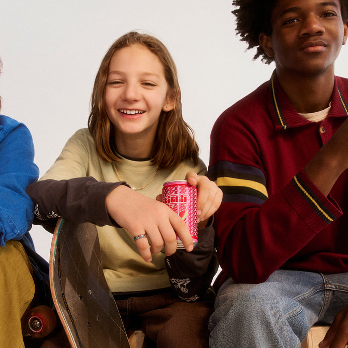 Three teens sitting together, one holding a red Stella Artois can, wearing casual colorful clothing with a skateboard visible