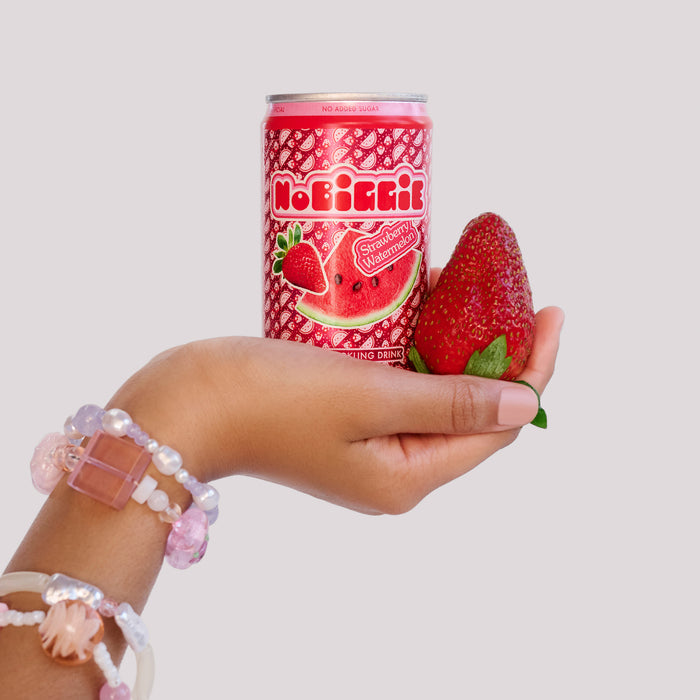 Hand holding NoBi&Kii strawberry watermelon drink can and fresh strawberry, wearing pink beaded bracelets against white background