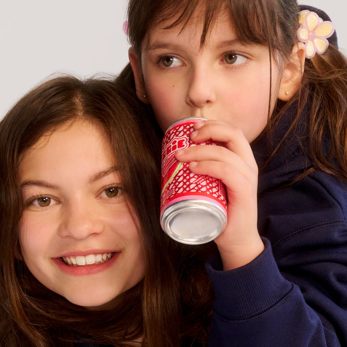 Two young girls sharing a red and white canned drink, one smiling at camera, other drinking from straw with flower hair clip