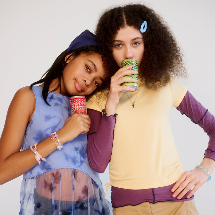 Two young women in colorful outfits drinking from decorative cups - one in blue tie-dye with hoodie, one in yellow top with curly hair and blue hair clip