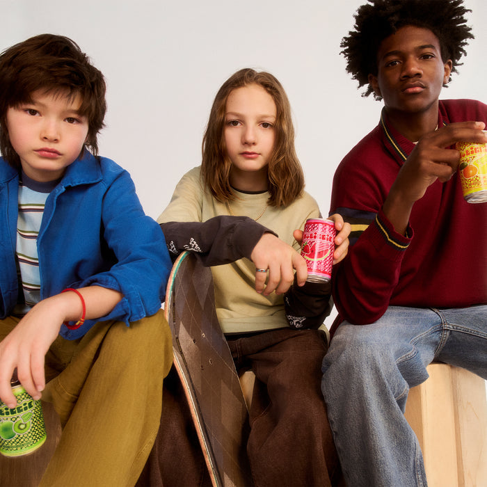Three teenagers sitting together holding colorful soda cans - one in blue jacket with green can, one in beige sweater with red can, and one in burgundy sweatshirt with yellow can.